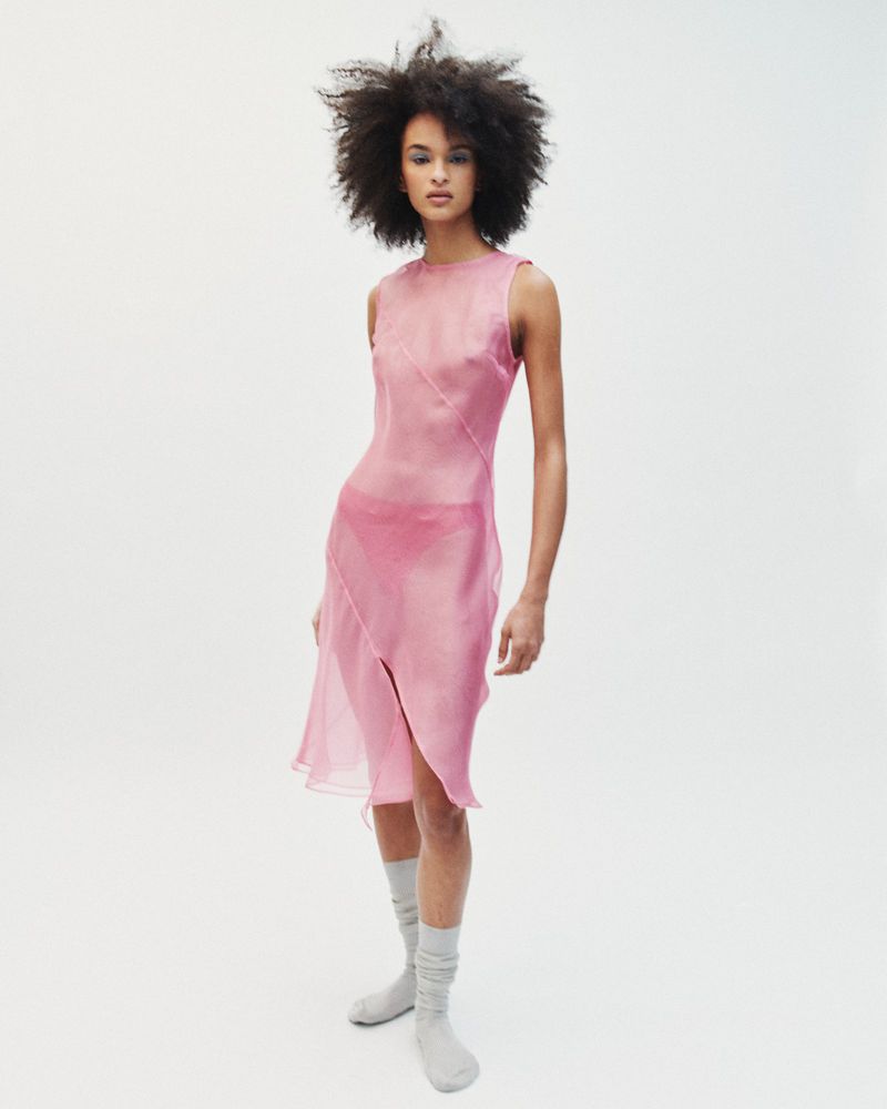 Model with voluminous dark hair wearing a sheer pink sleeveless dress and grey socks against a plain white background