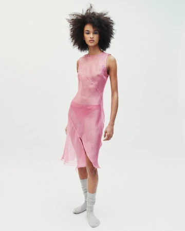 Model with voluminous dark hair wearing a sheer pink sleeveless dress and grey socks against a plain white background