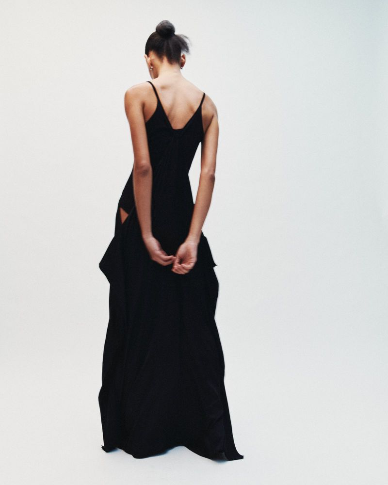 Model seen from behind wearing a long black dress with thin straps against a plain white background.