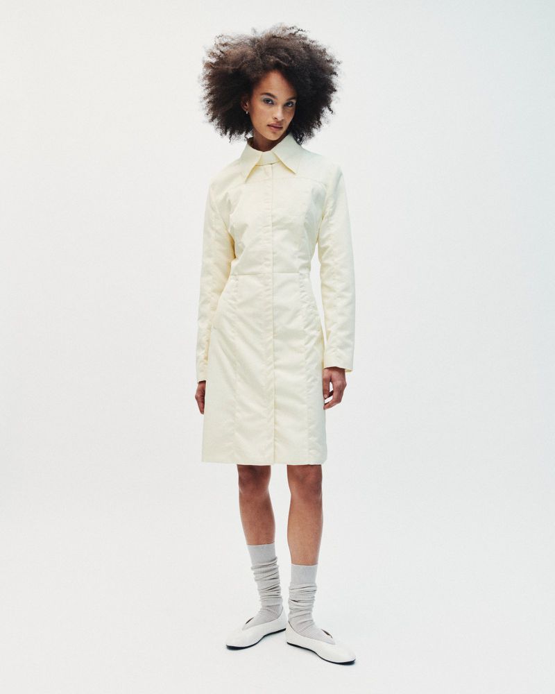 Model with voluminous dark hair wearing a light cream button-down coat and grey socks against a white seamless background.