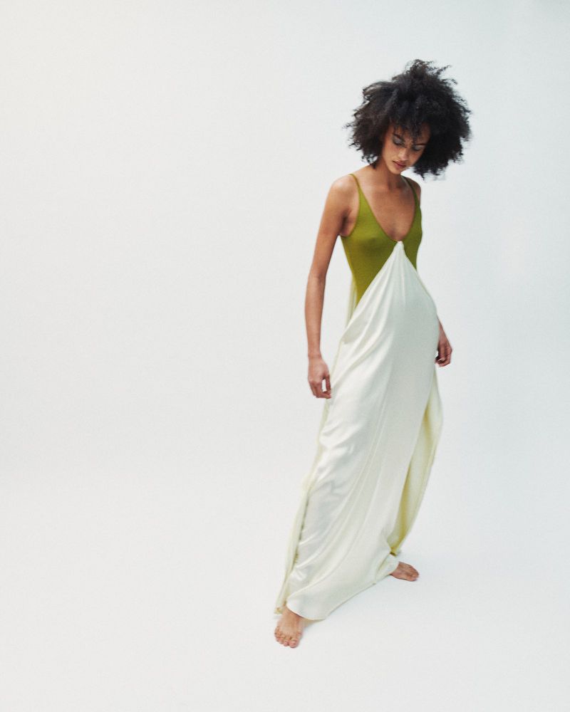 Model with dark curly hair wearing an olive green and white dress standing against white seamless background