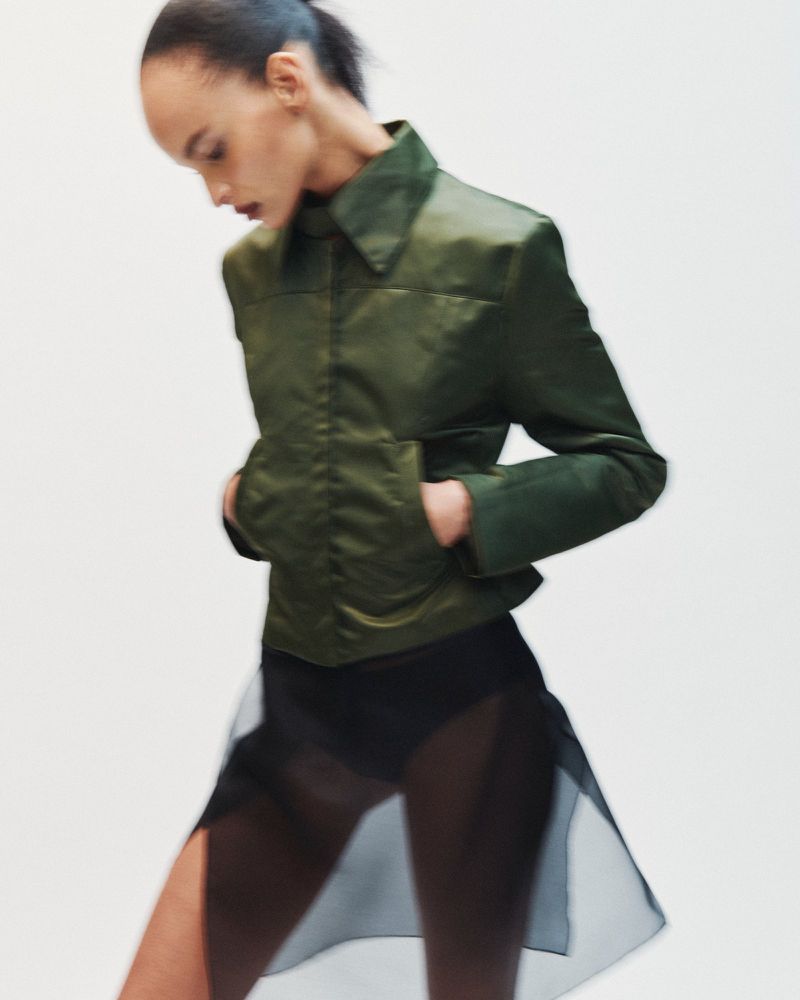 Model in motion wearing a structured green jacket and sheer black skirt against a plain white background