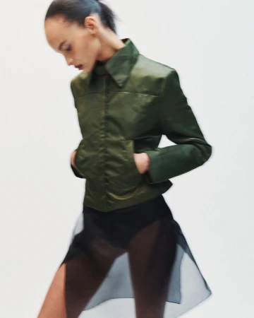 Model in motion wearing a structured green jacket and sheer black skirt against a plain white background