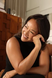 Portrait of Mary Phillips smiling with hand to cheek, wearing a black sleeveless top against an interior living room background.