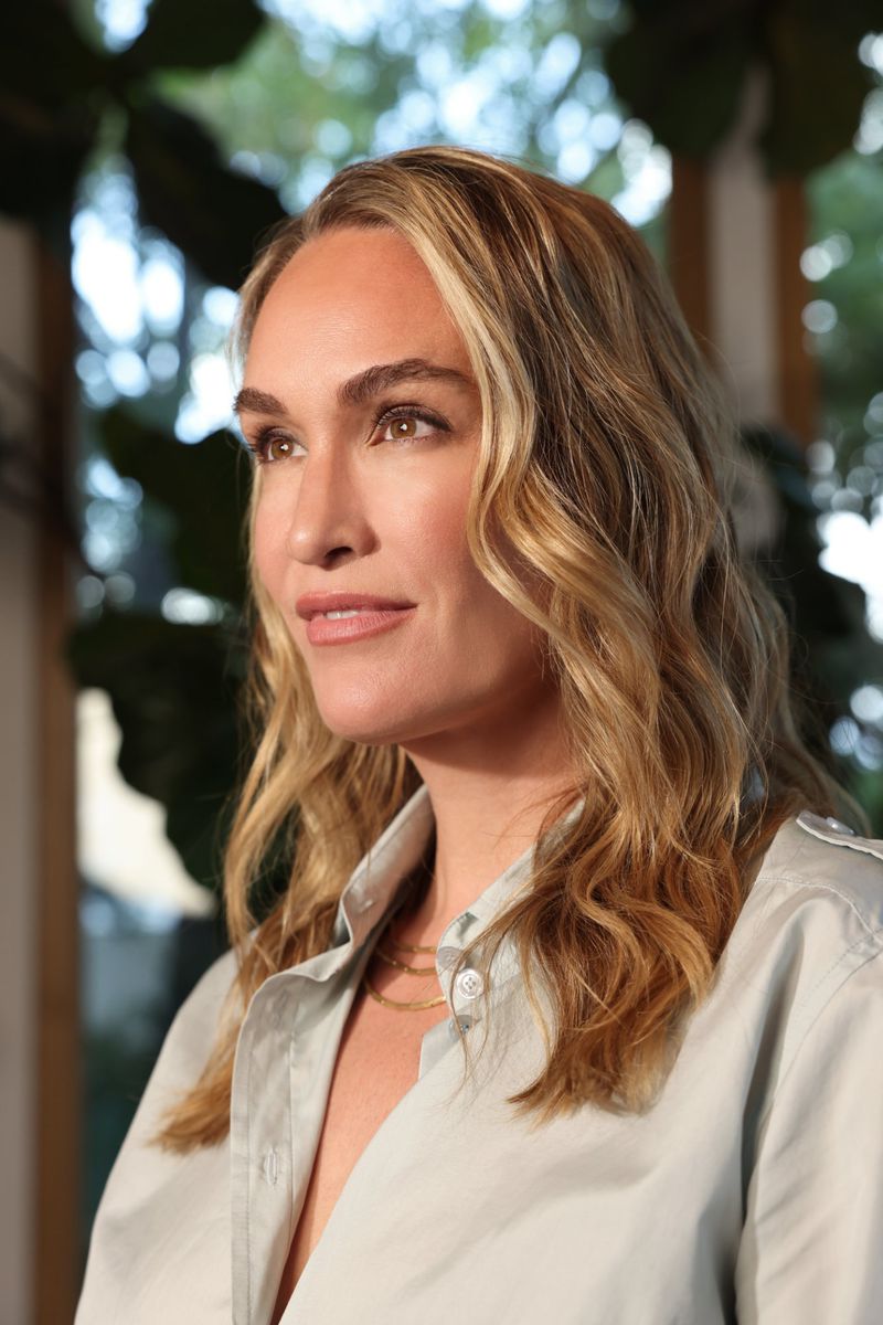 Close-up portrait of a woman with blonde highlighted hair wearing a light grey button-down shirt against an indoor background.