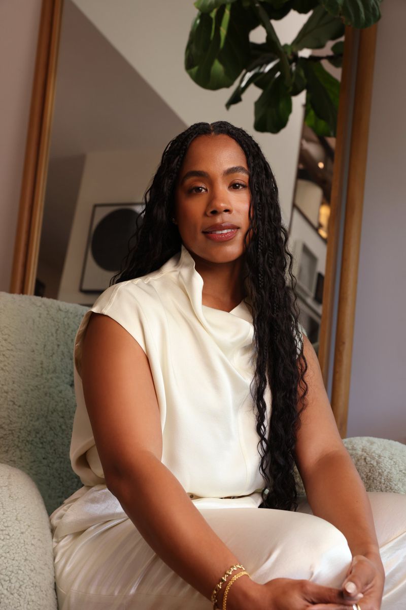 Portrait of a black woman with long dark hair wearing a white top seated in an armchair for La Mer.