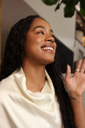 Close-up portrait of a smiling Black woman with long dark braided hair wearing a white satin top for La Mer.