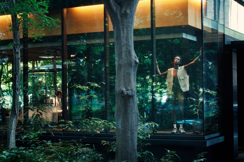 Model standing inside a modern glass pavilion wearing a utility vest with lush surrounding foliage and trees visible through the windows.