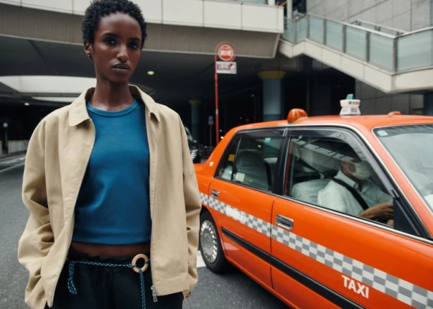 Model standing outdoors near an orange taxi wearing a tan jacket and blue top for Aman Essentials