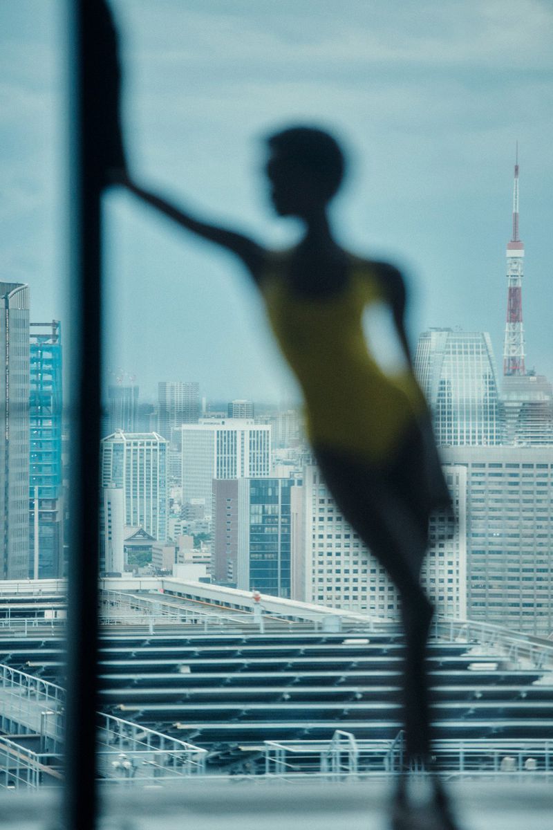 Silhouetted figure posed against a large window overlooking a city skyline with a tall red tower.