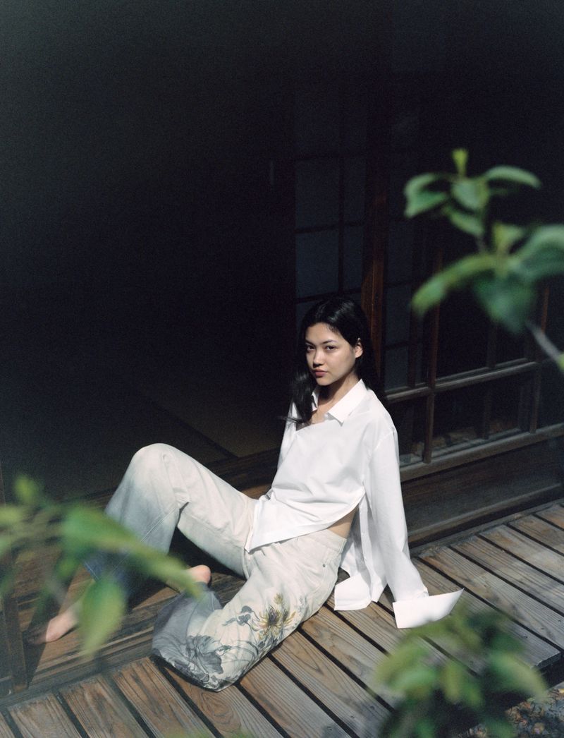 Asian woman in white shirt and patterned pants sits on a wooden porch in front of a dark doorway with green leaves in foreground.