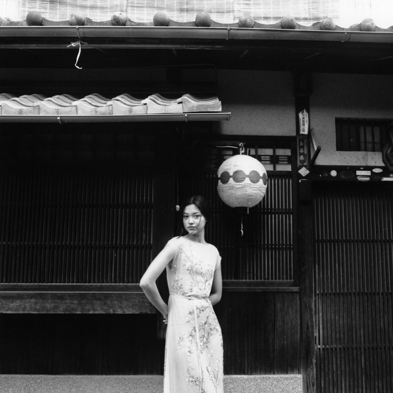 Woman in long dress poses in front of traditional Japanese building with lantern. Black and white photo with shallow depth of field.