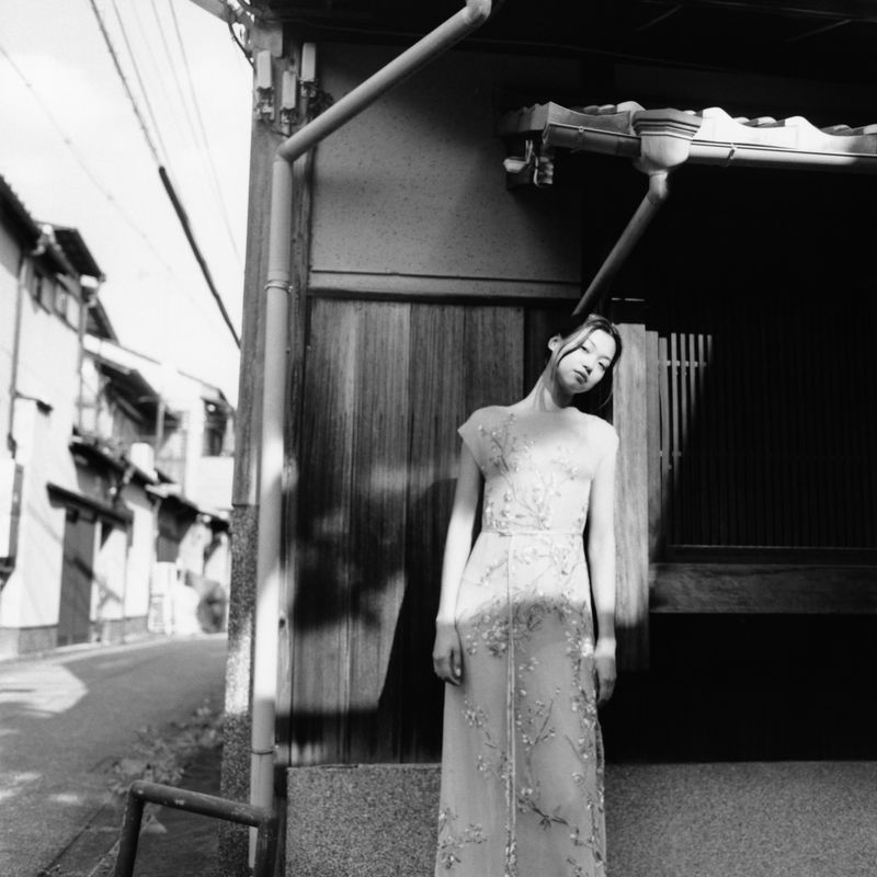 Monochrome portrait of a slim, young adult female in a light-colored dress, standing outside an old building on a narrow street in Japan.