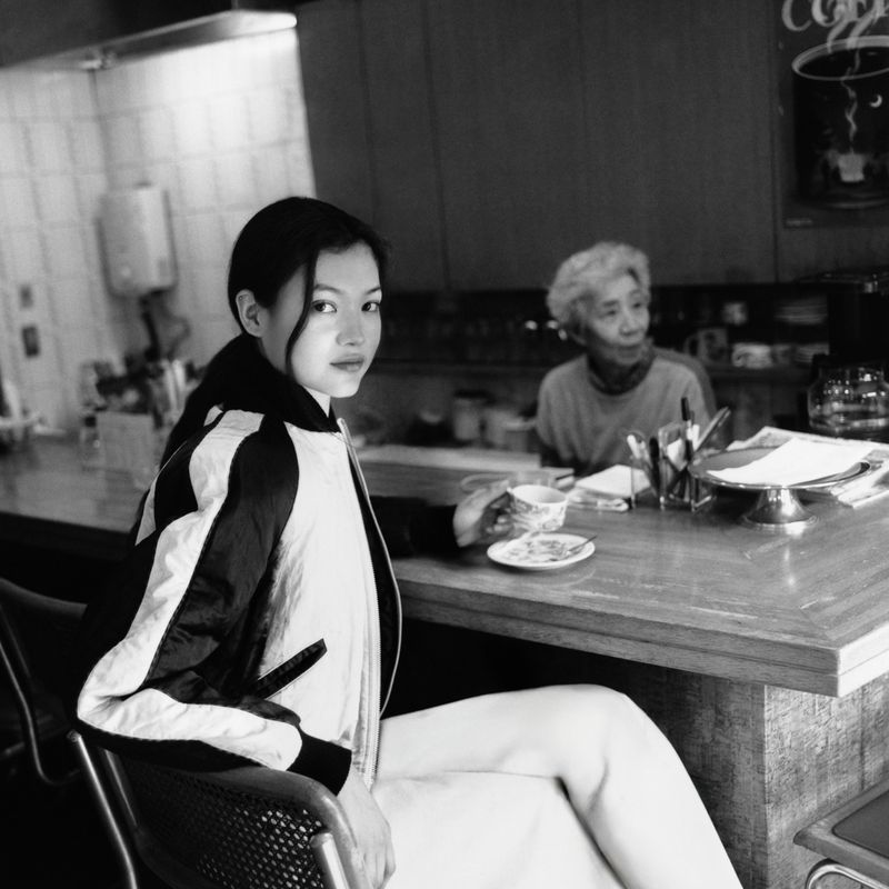 Black and white portrait of an Asian woman in jacket at a counter, an elderly woman sits behind her in a coffee shop.