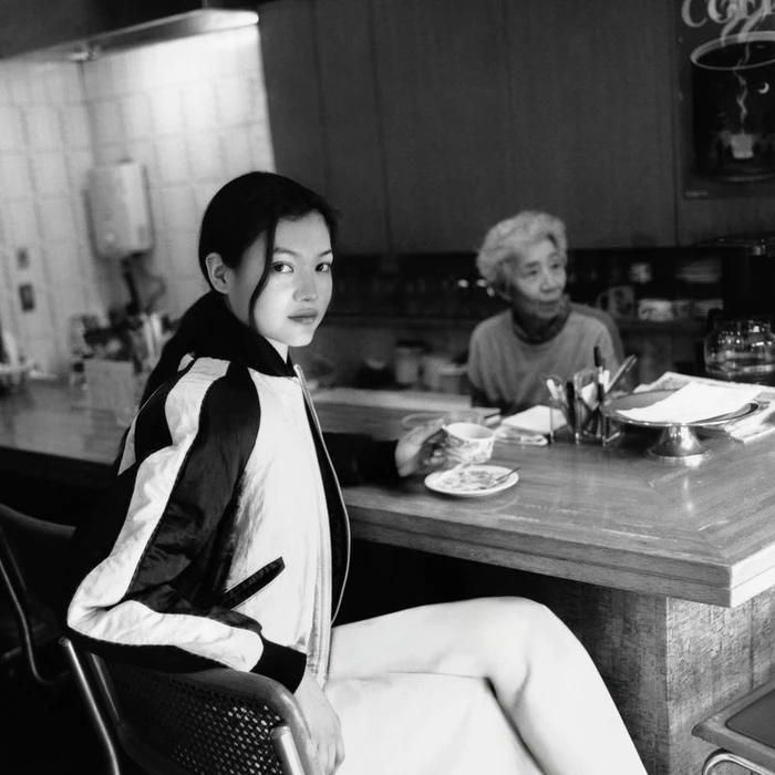 Black and white photo of model Mika Schneider sitting at a cafe counter holding a cup, with a woman standing behind the bar.
