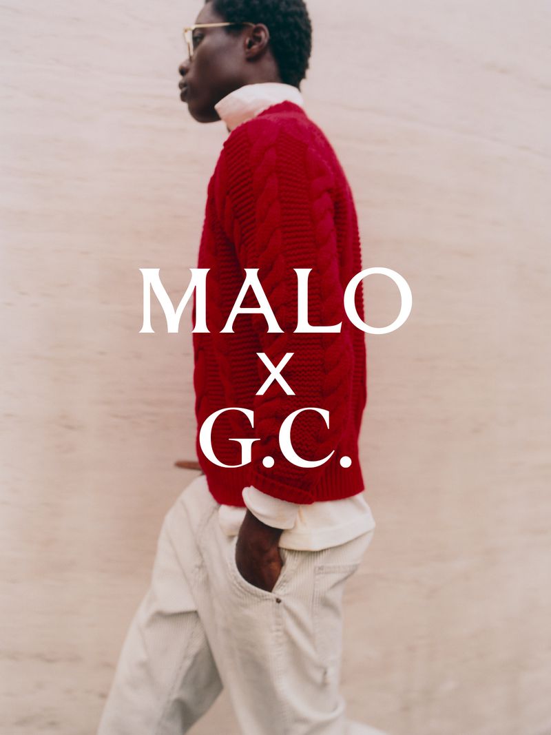 Profile view of a Black man wearing a red cable knit sweater and light trousers walking past a textured beige wall