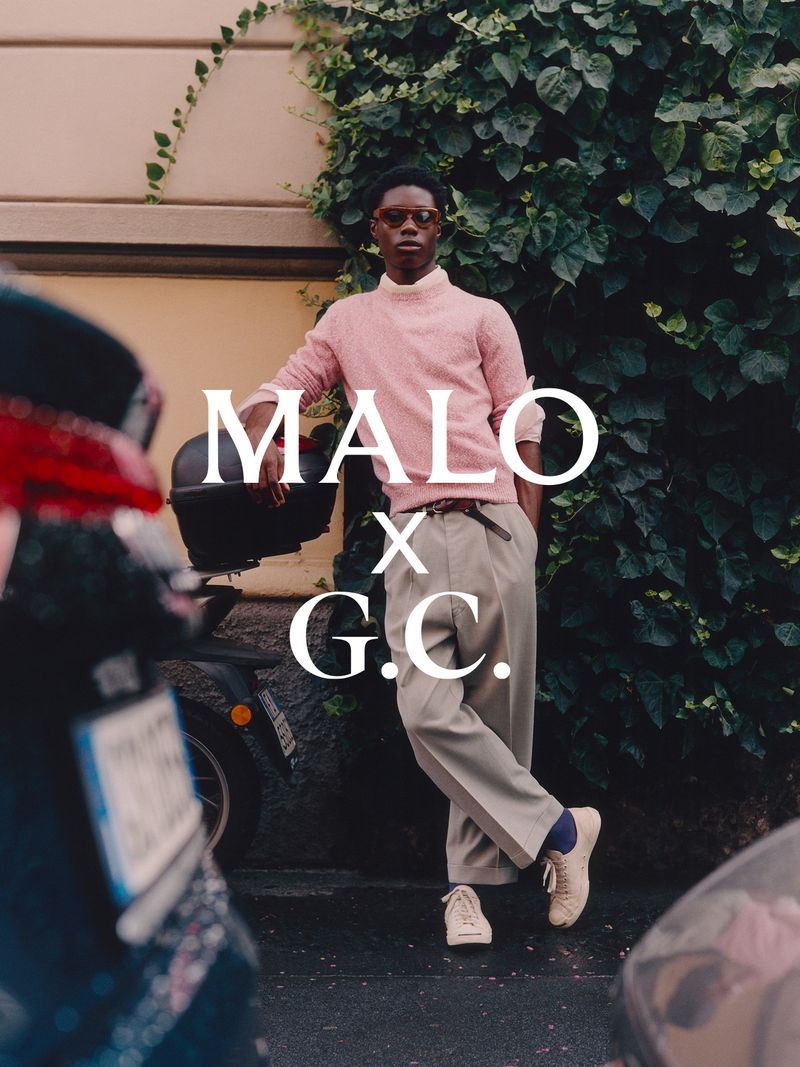 Black man wearing a pink knit sweater and beige trousers leaning against a wall covered in green ivy, photographed by Cornelius Kaess.