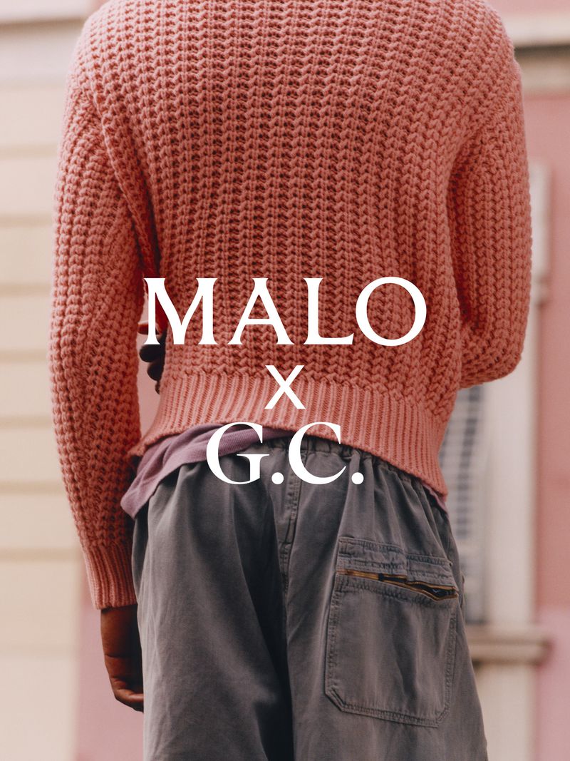 Rear view of a person wearing a pink textured knit sweater and grey trousers against a blurred building background
