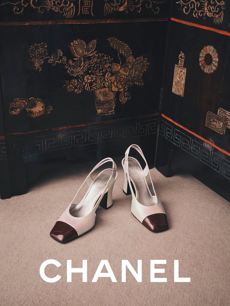 Two-tone slingback heels with dark cap toes placed on a beige carpet against a dark lacquered folding screen with gold floral motifs.