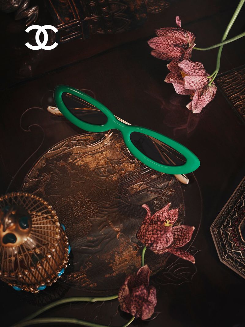 Green oval eyeglasses resting on an ornate dark surface with checkered flowers and a small decorative birdcage