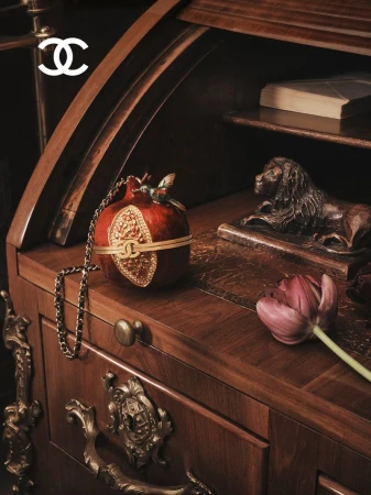 Pomegranate-shaped novelty handbag resting on a dark wood rolltop desk next to a bronze lion figurine and a flower.