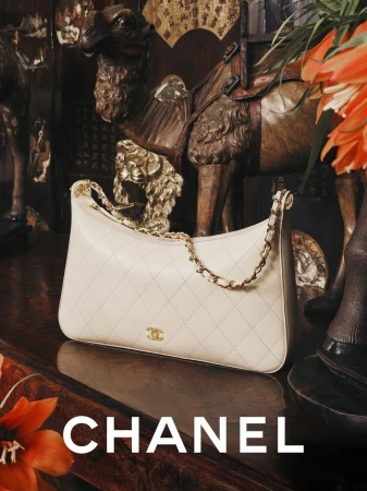 White quilted Chanel handbag with gold chain strap displayed on a dark wooden surface in front of an antique camel figurine
