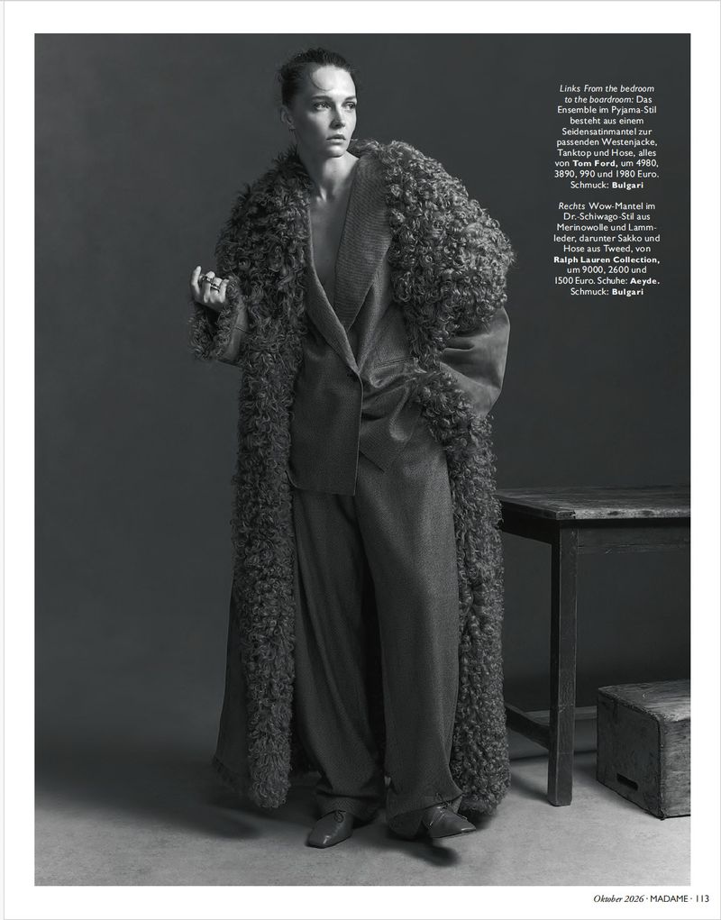 Model standing in a long textured fuzzy coat and tailored suit trousers against a grey studio backdrop.