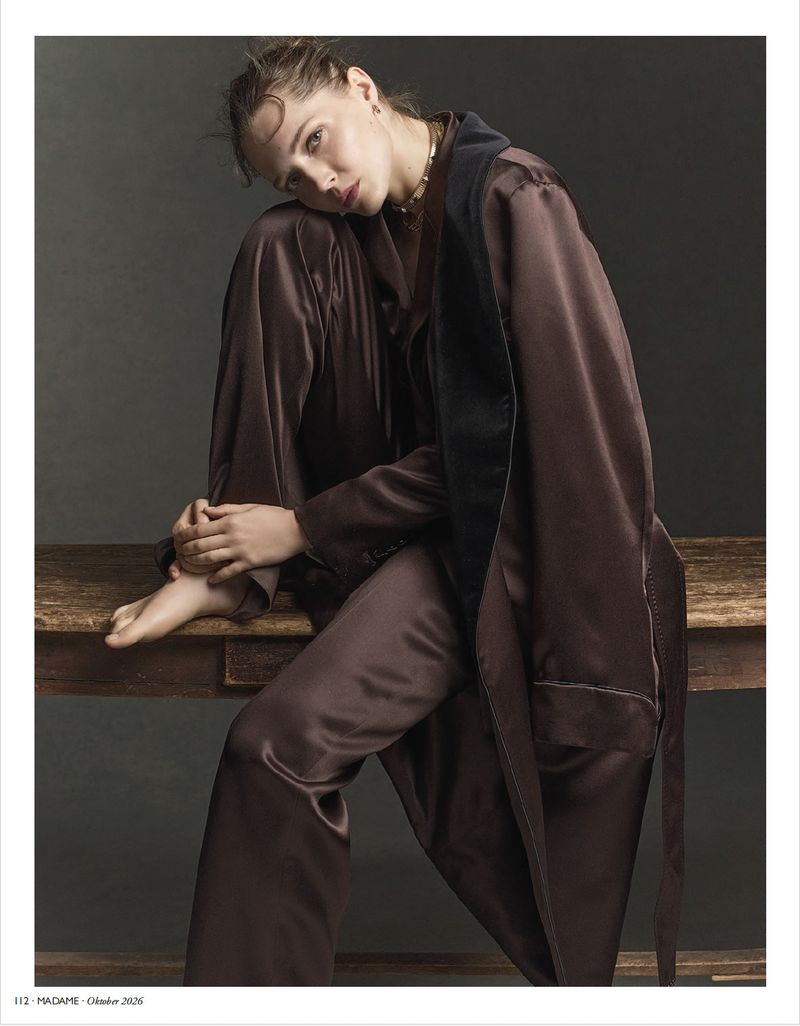 Woman in a brown silk pajama set and matching robe sitting on a wooden bench against a dark studio background