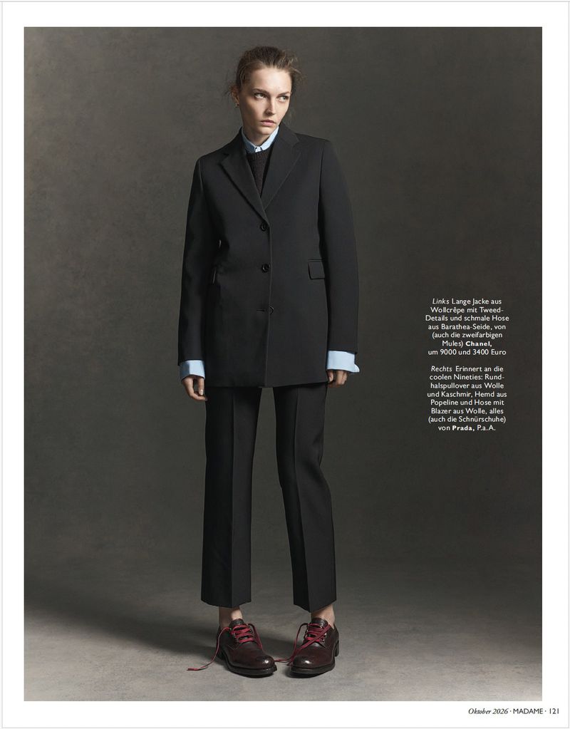 Model standing wearing a dark tailored suit jacket and trousers with dark shoes against a dark studio background