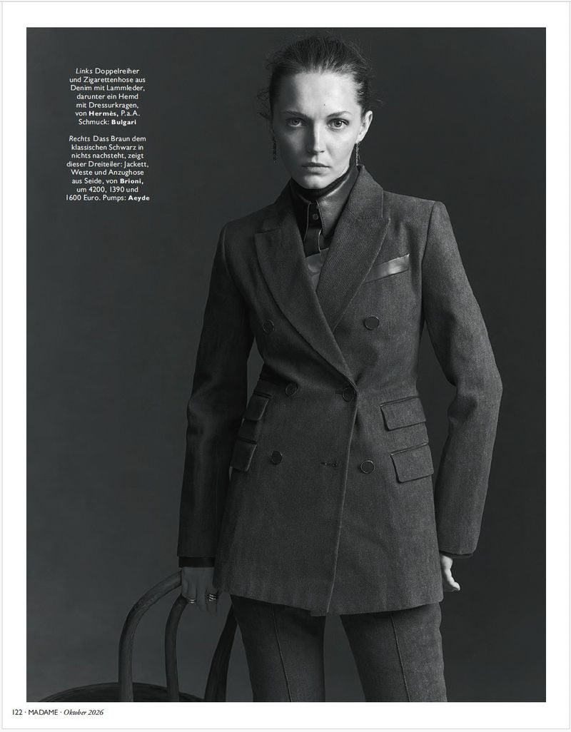 Black and white editorial portrait of a woman wearing a tailored double-breasted suit jacket against a dark studio background.