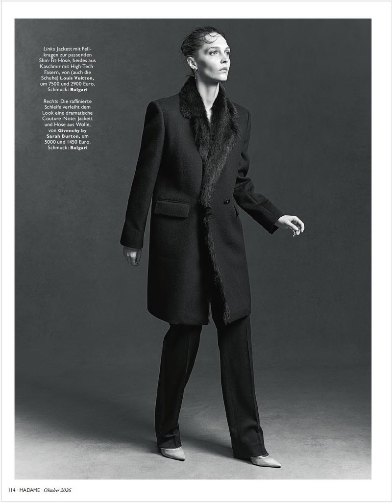 Model wearing a dark coat with fur collar and matching trousers against a grey seamless studio background.