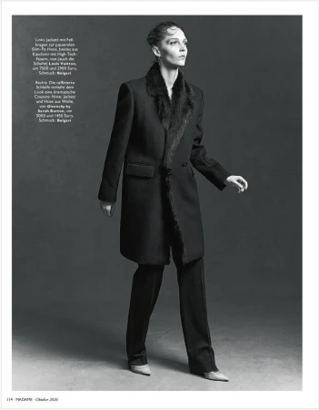 Model wearing a dark coat with fur collar and matching trousers against a grey seamless studio background.