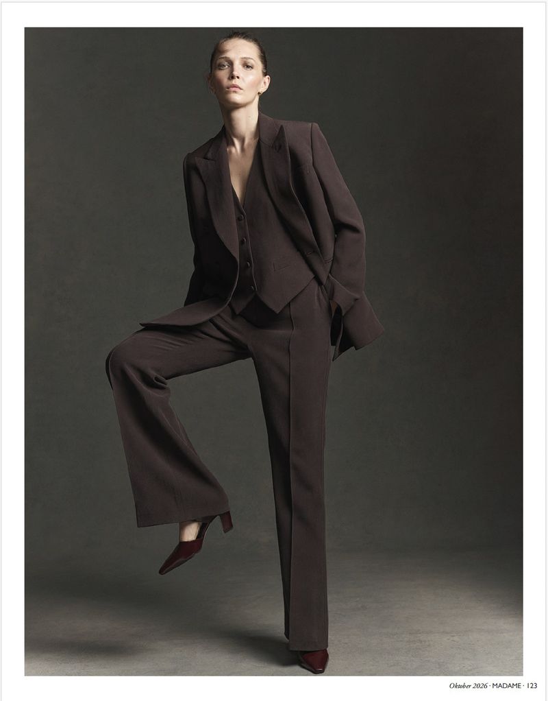 Model wearing a dark brown tailored suit standing with one leg raised against a dark grey studio background.