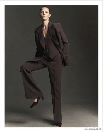 Model wearing a dark brown tailored suit standing with one leg raised against a dark grey studio background.