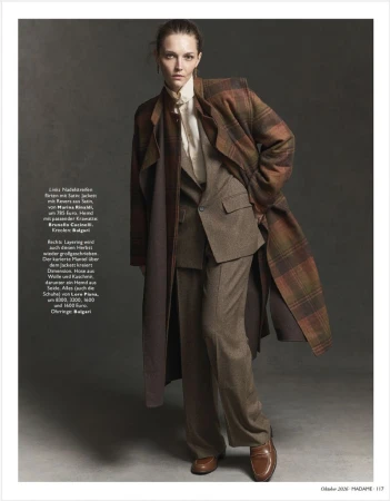 Model in layered brown suit and plaid coat standing against a dark grey studio background.
