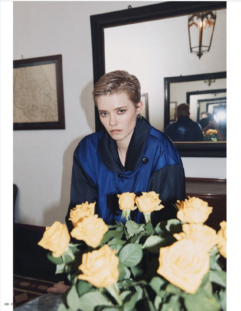 Model with short hair wearing a blue jacket behind yellow roses in a room with mirrors