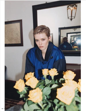 Model with short hair wearing a blue jacket behind yellow roses in a room with mirrors