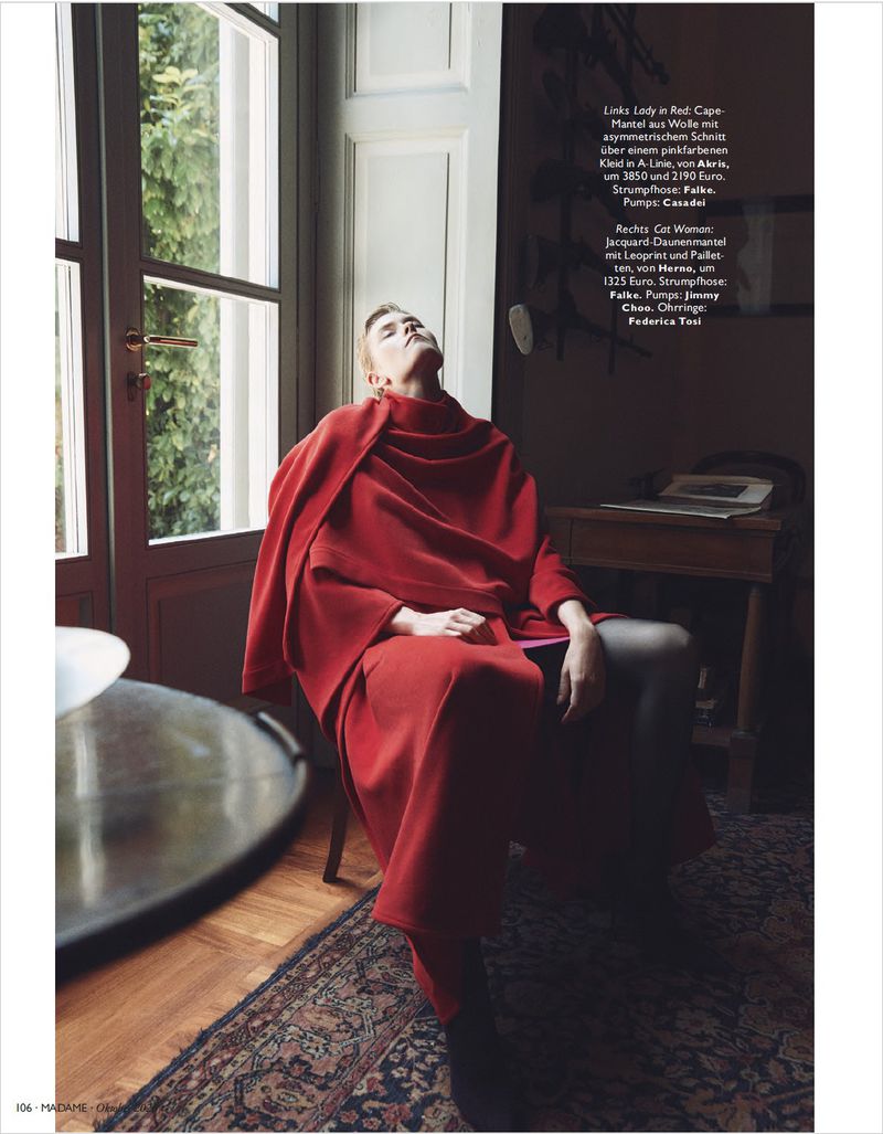 Model wearing a vibrant red wrap coat seated in an interior room with large windows, styled for Madame Germany.