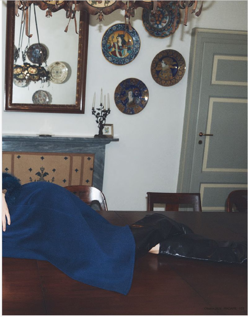 Model wearing a blue coat and black boots lying across a wooden dining table in a traditional interior