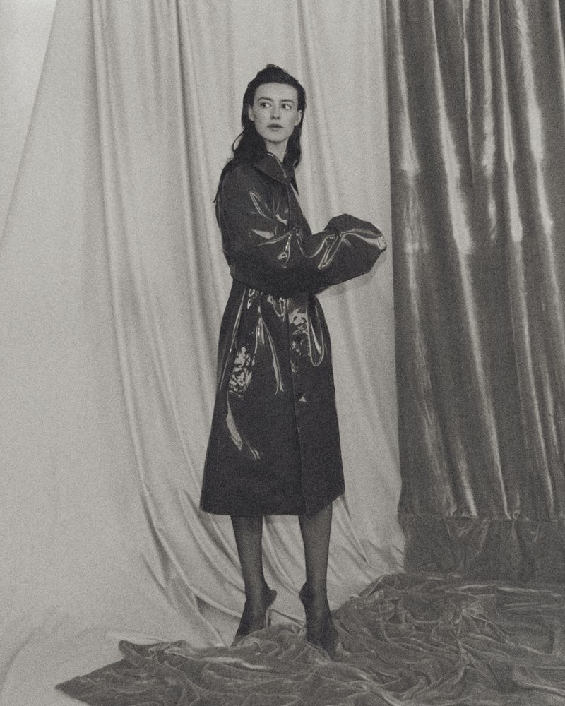 Portrait of actor Daisy Edgar-Jones in a glossy trench coat standing against draped fabric backdrops for Vogue España.