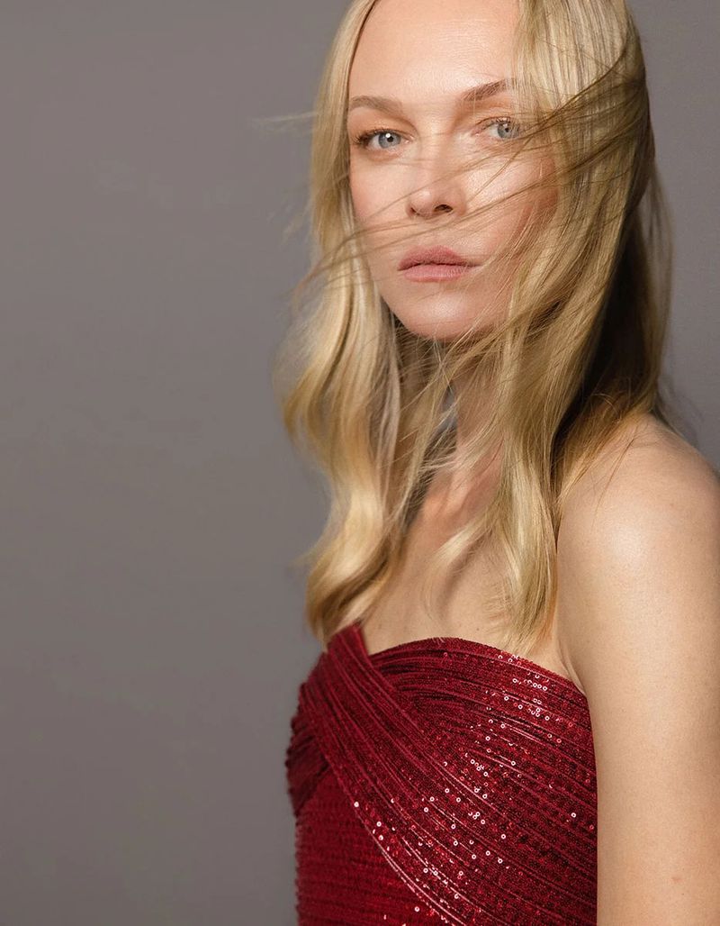 Portrait of a blonde woman wearing a strapless red sequin dress with wispy hair across her face against a grey backdrop.