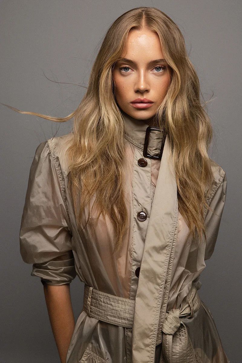 Portrait of a blonde woman wearing a belted beige trench coat against a solid grey background for Costume Norway.