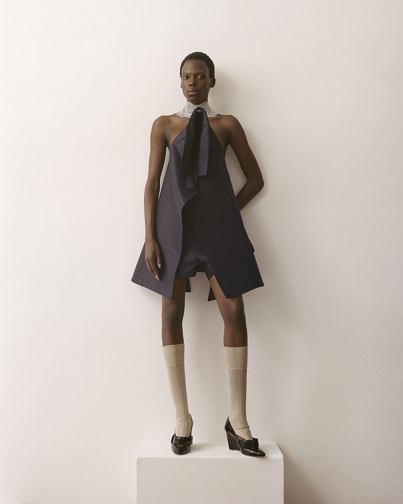 Model Agel Akol in a structured dark dress and knee-high socks standing on a white pedestal