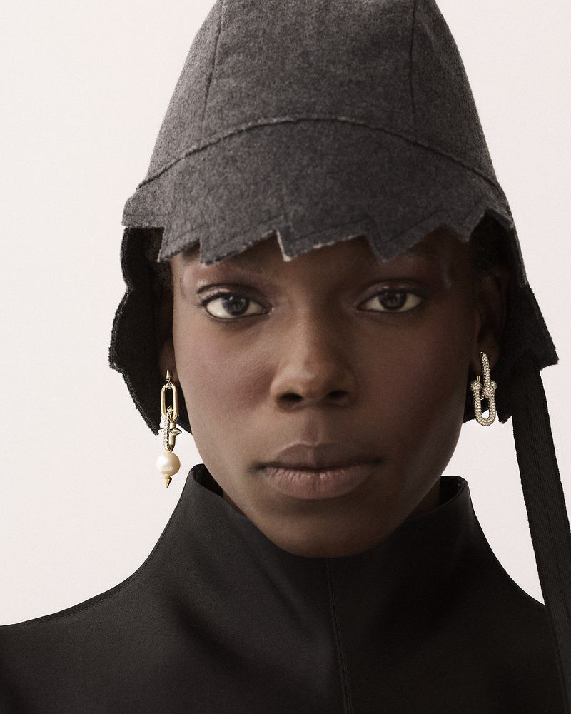 Close-up portrait of a model wearing a grey wool cap and dangling pearl earrings against a light background.