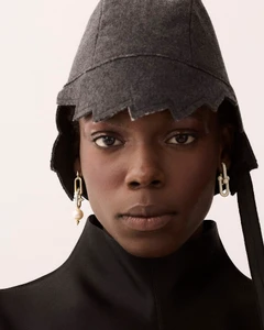 Close-up portrait of a model wearing a grey wool cap and dangling pearl earrings against a light background.