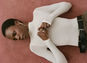 Model wearing diamond rings and a white long-sleeve top lying on a textured mauve surface for Heroine Magazine.