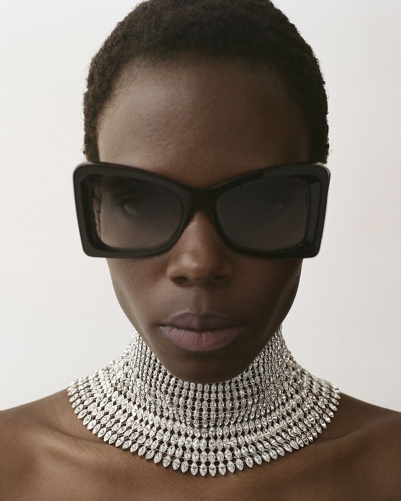 Close-up portrait of a model wearing large black sunglasses and a wide rhinestone choker necklace against a plain white background.