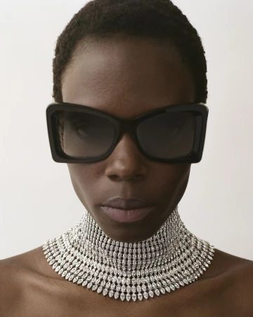 Close-up portrait of a model wearing large black sunglasses and a wide rhinestone choker necklace against a plain white background.