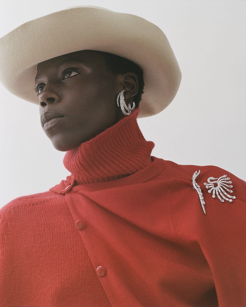 Model in red turtleneck and wide-brim hat wearing sparkling diamond earrings and brooches for Heroine Magazine.