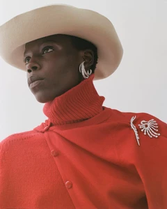 Model in red turtleneck and wide-brim hat wearing sparkling diamond earrings and brooches for Heroine Magazine.